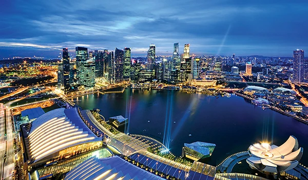 Vista aérea da orla de Marina Bay em Singapura ao entardecer, com arranha-céus iluminados e o icônico complexo de entretenimento The Merlion, refletindo nas águas da baía.