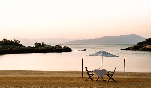 Foto da praia com uma mesa preparada para dois com guarda-sol, entre tochas.