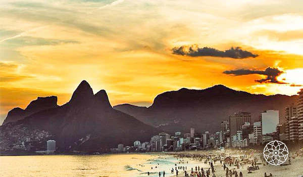 Paisagem da famosa praia de Ipanema ao entardecer, com o pôr do sol iluminando o céu dourado, o perfil das montanhas e o movimento das pessoas na areia.