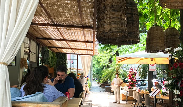 Imagem de um restaurante ao ar livre com clientes desfrutando de refeições sob sombra e árvores, decorado com luminárias de bambu e muitas plantas.
