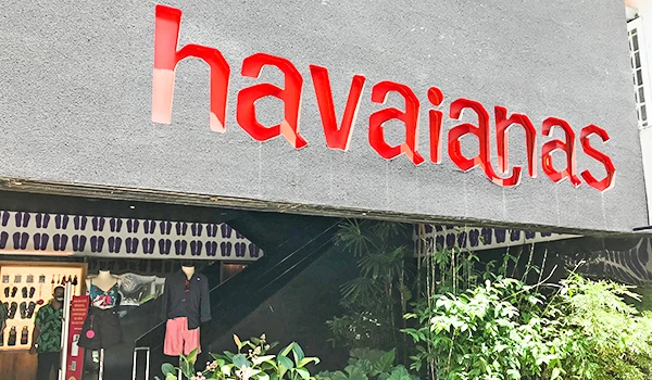 Fachada da loja havaianas com letreiro vermelho e entrada decorada com plantas, destaque para produtos de chinelos havaianas na vitrine