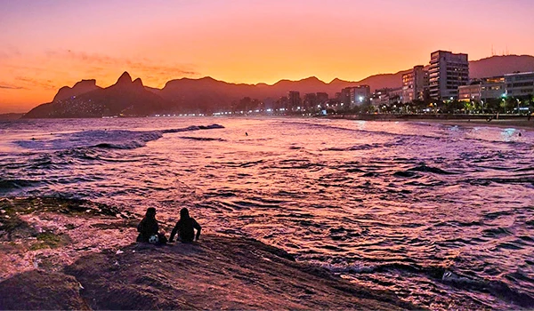 Pôr do sol na orla de Ipanema no Rio de Janeiro, com céu em tons de rosa e laranja, ondas suaves, dois pessoas se contemplando na areia, e skyline urbano ao fundo.