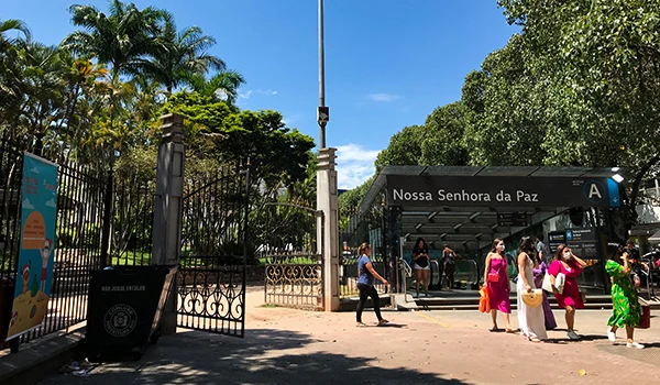 Entrada da Estação de Metrô em Ipanema com pessoas entrando e saindo, cercada por árvores e vegetação, dia ensolarado com céu azul.