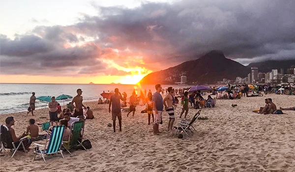 Turistas aproveitando o pôr do sol na praia com pessoas relaxando, cadeiras e guarda-sóis, cenário de montanha ao fundo, atmosfera de lazer e tranquilidade. Viagens para Ipanema.