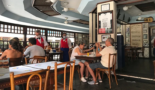 Clientes sorridentes e atendentes com máscaras em restaurante movimentado, aproveitando refeições com conforto.