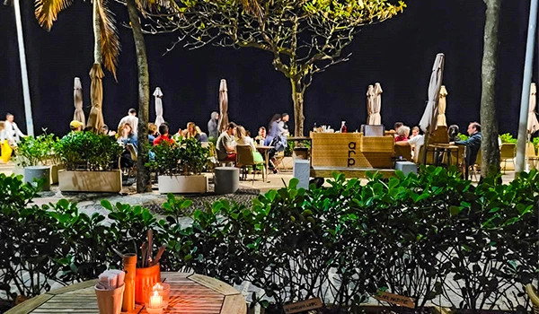 Imagem de um restaurante ao ar livre à noite, com mesas, cadeiras, guarda-sóis fechados e pessoas apreciando a experiência em um ambiente acolhedor e bem iluminado. Viagens para Ipanema.
