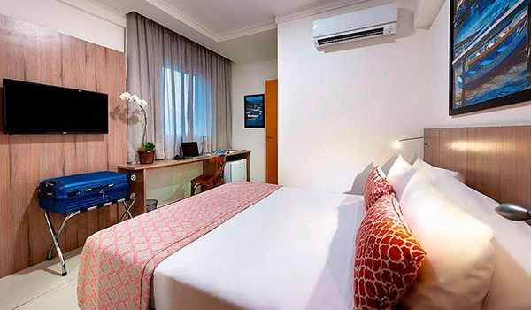 Imagem de um quarto de hotel moderno com cama confortável, TV na parede, cortinas largas, decoração sóbria e ar-condicionado, ideal para descanso e hospedagem. Lua de Mel em Cabo Frio.