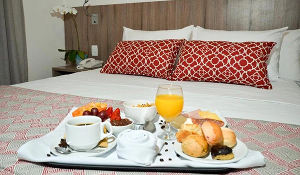 Café da manhã com pão, bolos, frutas, suco de laranja e café servido na cama de um hotel, perfeito para começar o dia de forma especial.