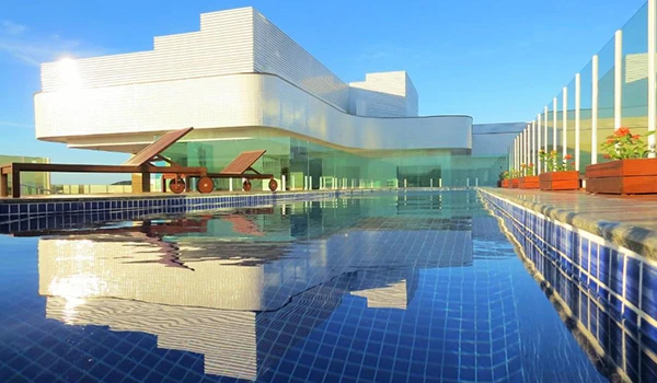 Imagem de uma piscina de borda infinita com vista para um prédio de arquitetura moderna e jardim, sob um céu azul claro. Ideal para relaxamento e lazer. Lua de Mel em Cabo Frio.