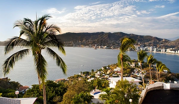Vista panorâmica de uma cidade costeira com árvores e palmeiras, vista para o mar e colinas ao fundo, céu parcialmente nublado em um dia ensolarado. Viagem ao México.