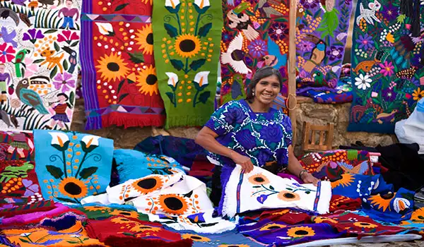 Mulher vendendo artesanato de bordado com telas coloridas e padrões florais tradicionais, em feira ao ar livre, destacando a cultura artesanal.mexicana.
