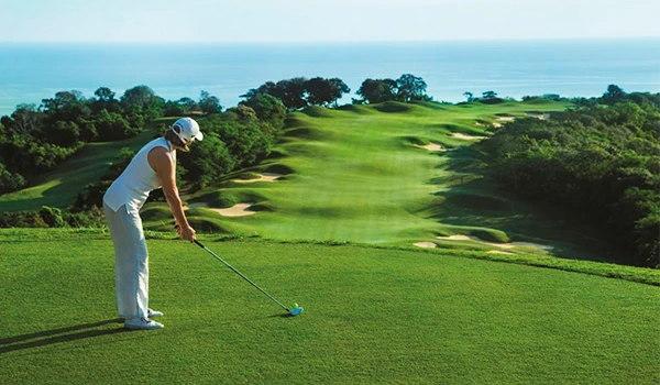 Jogadora de golfe se preparando para realizar um tiro em um campo de golfe bem cuidado com o mar ao fundo, árvores e areia na paisagem.Lua de Mel na Jamaica.