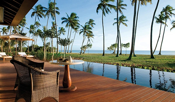 Vista relaxante de um deck com mesas e cadeiras, cercado por palmeiras e com vista para o mar calmo, ideal para momentos de tranquilidade e lazer.