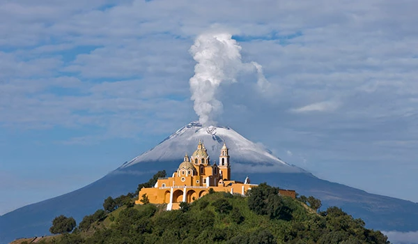 Vista panorâmica do vulcão de fumaça com uma igreja colorida na base, cercada de vegetação verde, sob céu azul com nuvens. Viagem ao México.
