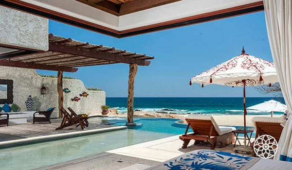 Imagem de uma varanda com vista para a praia, piscina, espreguiçadeiras de madeira, guarda-sol, cortinas brancas e um cenário oceânico ensolarado.