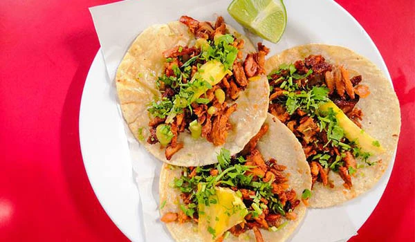 Três tacos de carne bovina com queijo, cebola, coentro, limão e molho, servidos em prato branco sobre fundo vermelho.
