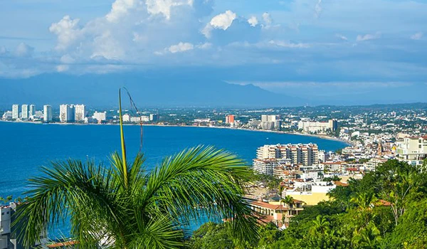 Vista panorâmica da cidade de Cancún com mar azul claro, arranha-céus modernos e vegetação tropical ao fundo, com céu parcialmente nublado e montanhas ao longe. Viagem ao México.