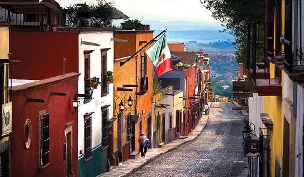 Cena de uma rua colorida na Viagem ao México, com casas de várias cores, ladeada por árvores e uma vista para o horizonte ao fundo. Pessoas caminhando na calçada, criando um ambiente turístico e acolhedor.