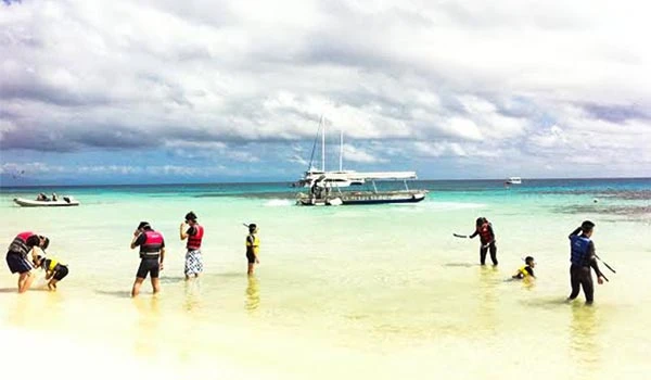 Turistas explorando a água cristalina da praia com um barco ao fundo, céu parcialmente nublado, ambiente de lazer e aventura na Austrália.