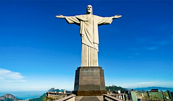 Vista da estátua do Cristo Redentor com braços abertos em frente à cidade do Rio de Janeiro, Brasil, sob céu claro e azul.