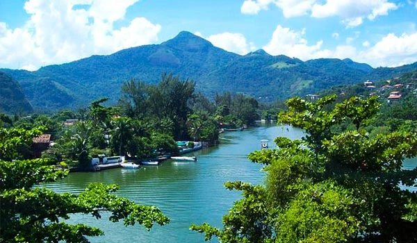 Vista panorâmica de um rio cercado por muita vegetação verde, com montanhas ao fundo e céu azul claro com algumas nuvens, ideal para atividades ao ar livre na natureza, uma das atrações da Barra da Tijuca.