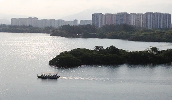 Vista panorâmica da Lagoa  de Marapendi - Barra da Tijuca - RJ, com prédios altos ao fundo e uma área verde na frente, sob um céu claro e com montanhas ao longe.