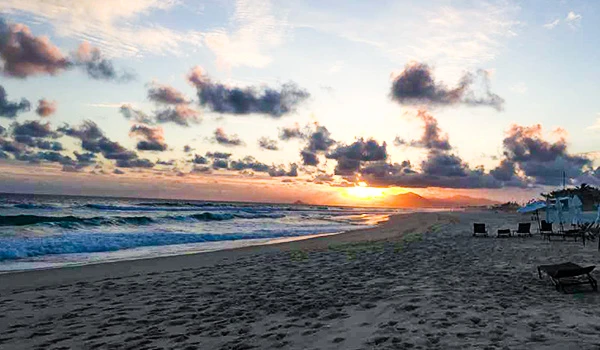 Pôr do sol na praia com nuvens no céu, ondas do mar e areia, criando uma cena tranquila e acolhedora. Perfeito para quem busca relaxamento à beira-mar.