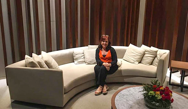 Deusa Rodrigues sentada em sofá branco em ambiente interno com decoração moderna de madeira e plantas, dicas sobre o que fazer Barra da Tijuca.