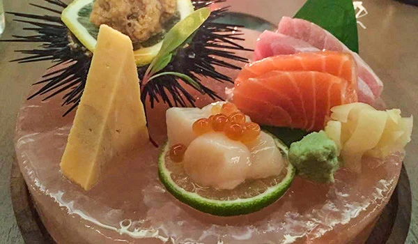 Prato de sashimi com fatias de salmão, atum e outros peixes, acompanhado de wasabi, gengibre, limão com caviar e uma decoração de folhas verdes, servido em uma bandeja de vidro.