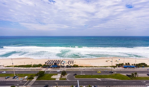 Praia de ondas fortes com areia clara, cabanas de praia e estacionamento ao fundo, sob céu parcialmente nublado, ideal para descanso e lazer à beira-mar. O que fazer na Barra da Tijuca.