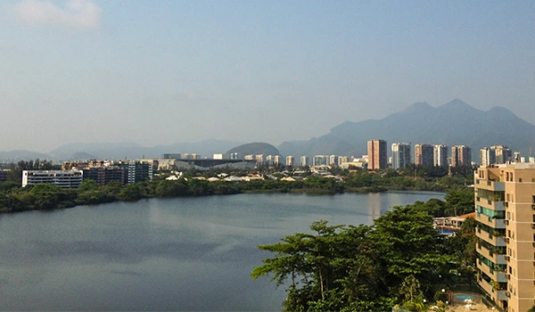 Vista panorâmica da Lagoa  de Marapendi - Barra da Tijuca - RJ, com prédios altos ao fundo e uma área verde na frente, sob um céu claro e com montanhas ao longe.
