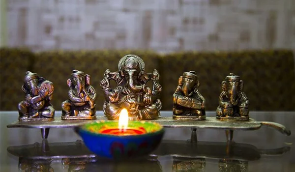 Estatuetas de divindades hindus, incluindo Ganesha, dispostas sobre uma prateleira, com uma lamparina de óleo colorida acesa em primeiro plano, em um ambiente decorado para o Diwali.