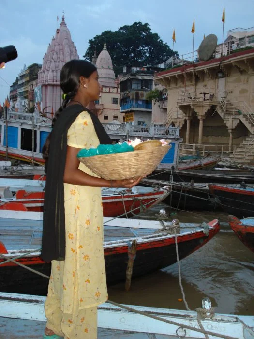 Mulher em pé, usando vestido amarelo florido e xale preto, carregando uma cesta de alimentos. Ao fundo, há barcos coloridos atracados às margens de um rio, edifícios e templos de arquitetura indiana.