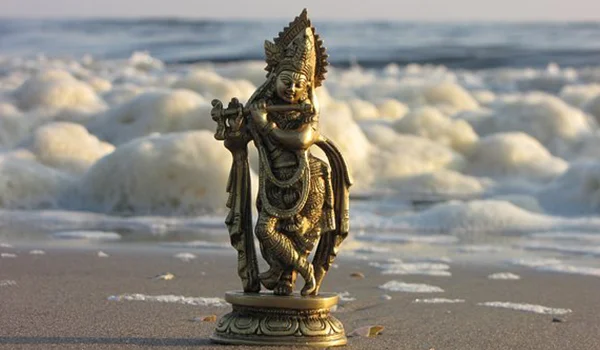 Estátua de uma divindade hindu tocando flauta, posicionada sobre um pedestal na areia de uma praia, diante de ondas do mar e espuma branca. A luz suave destaca os detalhes ornamentais da estátua.