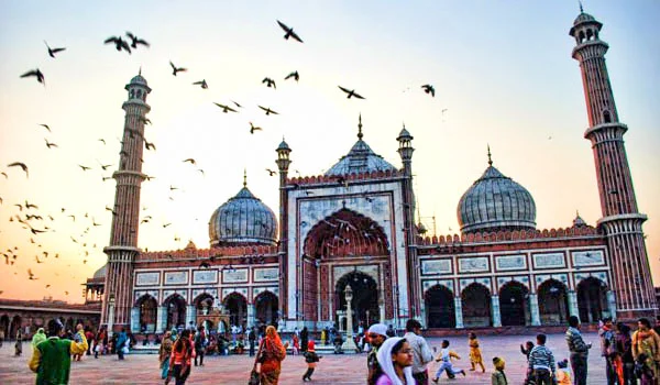 Mesquita Jama Masjid, em Nova Délhi, Índia, com suas cúpulas e minaretes, cercada por pessoas e muitas aves voando no céu.