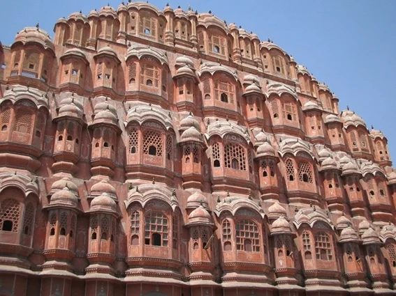 Fachada ornamentada do Hawa Mahal, em Jaipur, Índia, construída em arenito rosa, com várias janelas e sacadas de treliça finamente trabalhadas. O céu azul aparece ao fundo.