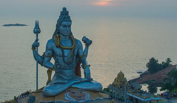 Estátua monumental azul do deus hindu Shiva, sentado em posição meditativa diante do mar, segurando um tridente. Ao fundo, há o oceano, pequenas ilhas e parte de um templo ou complexo religioso costeiro.