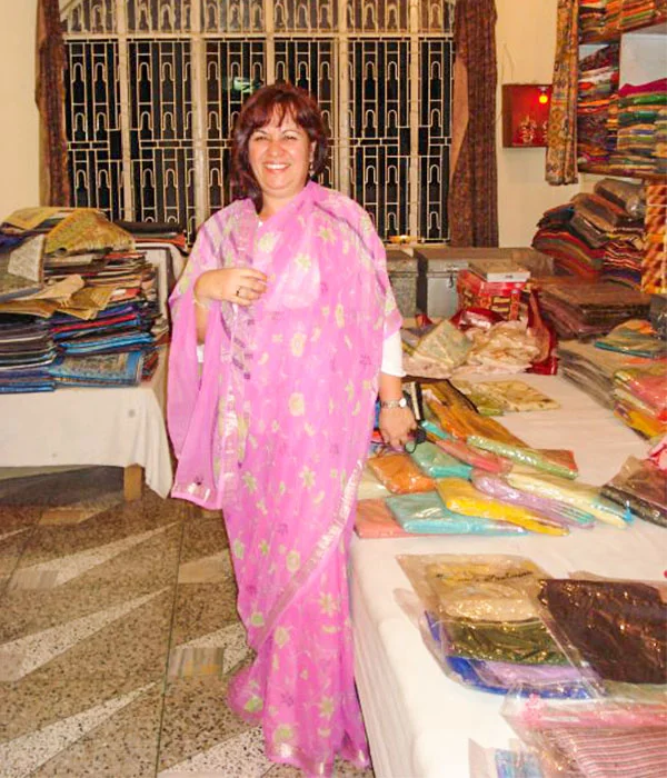 Deusa Rodrigues em uma loja de tecidos, usando um traje tradicional indiano rosa com detalhes florais. Ao redor dela há mesas e prateleiras repletas de tecidos coloridos dobrados e embalados. Ao fundo, vê-se uma grande janela com grades decorativas.