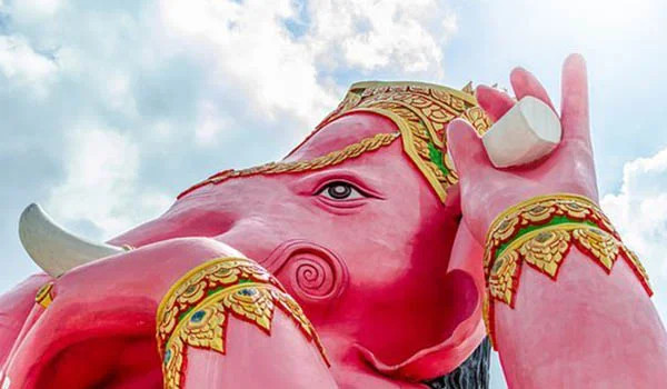 Estátua de uma divindade hindu com cabeça de elefante, representada em tom rosa vibrante e adornada com detalhes dourados e coloridos. A figura ergue uma das mãos, enquanto o céu azul com nuvens aparece ao fundo.