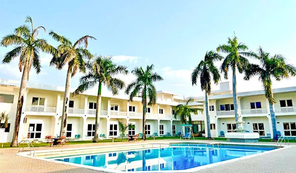 Hotel de dois andares cercado por palmeiras, com fachada branca, varandas e grandes janelas, voltado para uma piscina retangular azul em um pátio ensolarado.