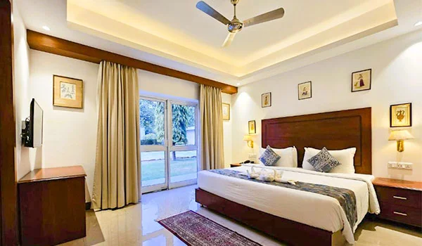 Quarto de hotel moderno e confortável, com cama de casal, cabeceira de madeira escura, roupas de cama brancas, mesas de cabeceira, TV na parede e portas de vidro que dão acesso a uma área externa ajardinada.