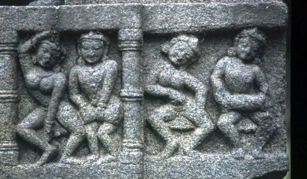 Baixo-relevo em pedra mostrando quatro figuras humanas sentadas ou agachadas, dispostas em dois grupos, separadas por uma coluna central. As figuras usam poucas vestimentas e apresentam posturas variadas, esculpidas em estilo antigo.