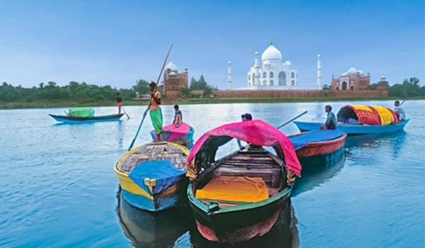 Barcos coloridos navegam sobre o rio Yamuna diante do Taj Mahal, na Índia, sob um céu azul. Pessoas estão nos barcos, incluindo uma embarcação coberta com tecido rosa e vermelho, enquanto o Taj Mahal branco se destaca ao fundo.