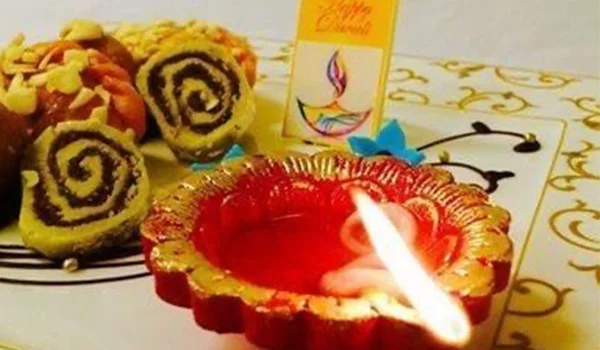 Imagem de uma decoração festiva com uma lamparina (diya) vermelha acesa em primeiro plano, doces enrolados ao fundo e um pequeno cartão decorativo sobre a mesa.