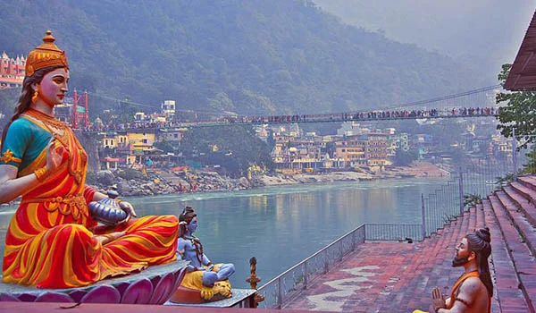 Estátua colorida de uma divindade hindu sentada em posição de meditação às margens de um rio. Ao fundo, há um templo e edifícios densamente agrupados junto às montanhas, além de uma ponte suspensa movimentada atravessando o rio. Na margem direita, uma pessoa sentada em postura de oração contempla a estátua. A paisagem montanhosa é parcialmente coberta por vegetação e névoa.