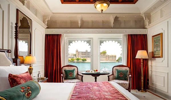 Quarto de luxo decorado em estilo tradicional indiano, com cama de casal, poltronas e cortinas vermelhas, janelas em arco com vista para um palácio à beira do lago e detalhes ornamentais nas paredes e no teto.