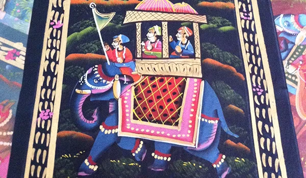 Tapete decorativo colorido com uma ilustração de um elefante azul ricamente ornamentado, carregando uma pequena estrutura semelhante a um palanquim com três pessoas em seu interior. O elefante usa adornos vermelhos, brancos e dourados, e ao fundo há árvores e vegetação em tons escuros. As bordas do tapete apresentam padrões decorativos florais e geométricos.