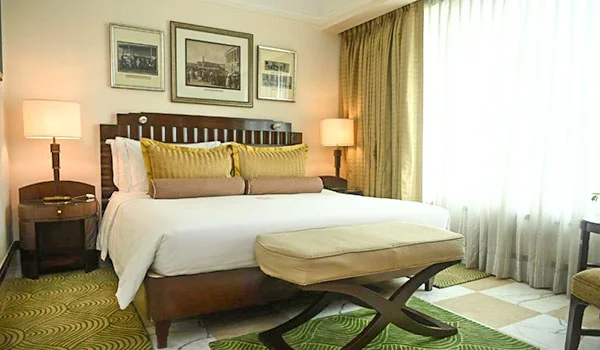Quarto de hotel elegante, com cama de casal de madeira escura, roupa de cama branca e almofadas em tons de amarelo e bege. Há mesas de cabeceira com luminárias, um banco estofado aos pés da cama e grandes janelas com cortinas claras.