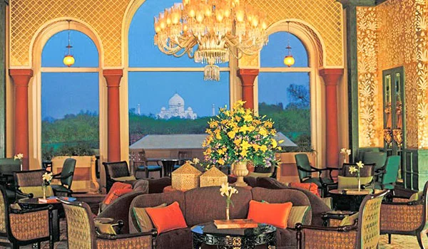 Salão elegante de estilo indiano, com lustre de cristal, grandes janelas em arco e poltronas ao redor de mesas baixas. Ao fundo, entre árvores, vê-se o Taj Mahal sob um céu azul.