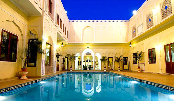Pátio interno de um palácio com piscina retangular de águas azuis, cercado por edifícios de arquitetura tradicional indiana, com arcos, janelas ornamentadas, palmeiras e iluminação quente.